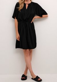 Svart kortärmad klänning med nyckelhålsutsnitt och dragsko i midjan, tillverkad av ett mjukt tyg, tillsammans med svarta platta sandaler.