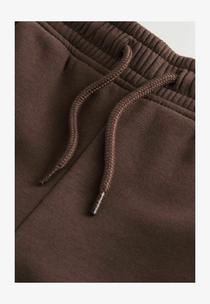 Pruunid puuvillased treeningpüksid sileda tekstuuriga, mis omavad ribaga vööd ja sobivaid nööre reguleeritava istuvuse jaoks.