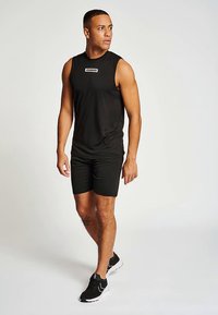 Man i svart ärmlös topp, svarta shorts och svarta sneakers som står och tittar åt vänster mot en enfärgad bakgrund.