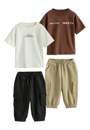 Barn iført olivengrønne cargobukser og hvide New Balance-sneakers står på et lyst gulv med en beige baggrund.
