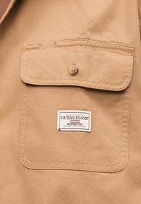 Gros plan sur une poche en tissu beige avec un rabat boutonné et une étiquette blanche Guess Jeans cousue sous le rabat.