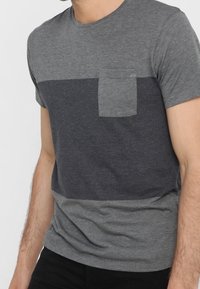 Camiseta gris con un diseño de bloques de color y un bolsillo en el pecho. La parte superior es de color gris oscuro, la parte inferior es de un gris más claro, hecha de una tela suave.