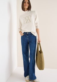 Světle béžová mikina s grafikou "COZY", spárovaná s tmavě modrými zvonovými džíny. Zelená olivová taška a bílé tenisky dokončují outfit.