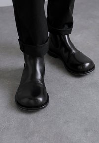 Bocanci negri din piele la glezna, cu vârful rotund, textură netedă, cusături minime și un profil scăzut, asociați cu pantaloni negri răsuciți.