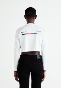 Fehér, rövidített, hosszú ujjú póló fekete kiegészítőkkel, amelyekben a "PEPE JEANS" és a "Red Bull Racing" logók találhatók. Fekete farmernadrággal párosítva.