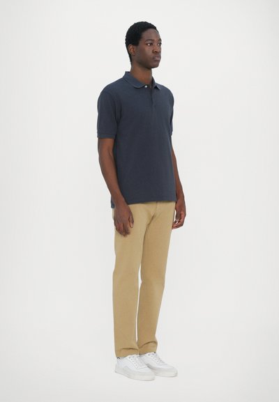 Σκούρο ναυτικό μπλουζάκι πόλο, με κοντά μανίκια, σχέδιο με γιακά. Καφέ παντελόνι, ίσια γραμμή. Λευκά αθλητικά παπούτσια με κορδόνια, επίπεδη σόλα.