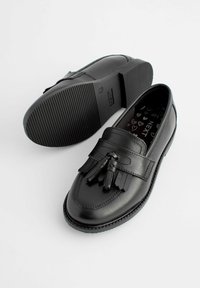 Mocasines de cuero negro con una correa decorativa con flecos y dos borlas. Suela de goma texturizada con una plantilla con diseño.