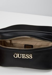 Černá kožená taška s zlatým logem "GUESS", se zipovým uzávěrem a béžovým interiérem s dalším zipovým kapsou. Hladká textura.