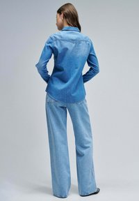 Light blue denim shirt with a collared neckline and long sleeves, featuring a curved hem. Paired with wide-leg light blue jeans.