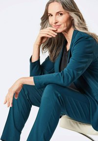 Teal pak met een getailleerde blazer en taps toelopende broek. Gladde stof, subtiele textuur en minimalistisch ontwerp. Model zittend tegen een neutrale achtergrond.