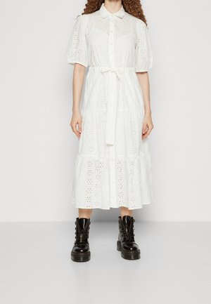 Femme portant une robe midi blanche à œillets avec des manches bouffantes et des bottines noires à lacets, debout devant un fond clair uni.