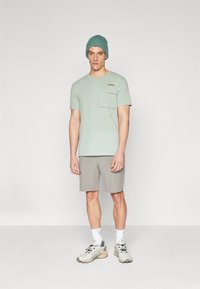 Ljusgrön bomulls-t-shirt med korta ärmar och bröstficka, i kombination med grå shorts och ljusa sneakers. Mössa i matchande grön.