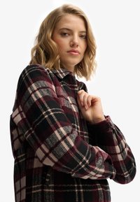 Koszula w kratę w odcieniach czerni, burgundu i beżu. Flanelowa tekstura, kołnierz i zapięcie na guziki. Modelka pozująca z założonymi rękoma.