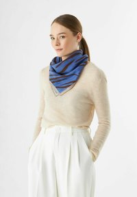 Blauwe sjaal met bruine strepen, gemaakt van zacht materiaal, gevouwen in een driehoekige vorm, gedragen over een lichtbeige sweater. Witte geplooide broek.