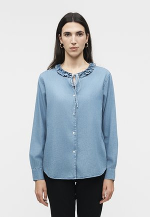 Γυναίκα με μακριά σκούρα μαλλιά φοράει ένα ανοιχτό γαλάζιο πουκάμισο με μακριά μανίκια και κουμπιά, με ντεκολτέ με βολάν και μαύρο παντελόνι, σε λευκό φόντο.