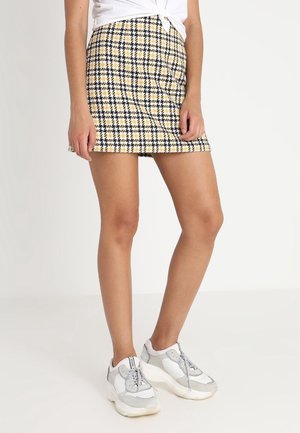 Frau trägt einen gelben, schwarzen und weißen karierten Minirock mit weißen und grauen Sneakers und einem weißen, geknoteten Top.