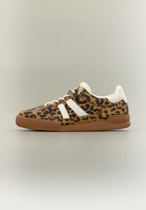 Alacsony szárú sportcipő barna és fekete leopárdmintával, fehér sarokkal és csíkokkal, fehér fűzőkkel, barna gumitalppal.