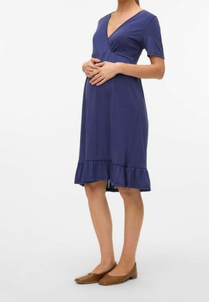 Kobieta w ciąży ubrana w granatową sukienkę do kolan z falbaną na dole oraz brązowe baleriny, delikatnie opierająca ręce na brzuchu.