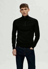 Sweater preto com fecho de pescoço feito de um tecido suave, com mangas longas, design justo e detalhes de costura mínimos, combinado com jeans escuros.