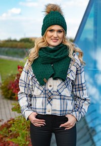 Beanie in maglia verde con pompon in pelliccia sintetica, sciarpa verde abbinata, giacca a bottoni a quadri beige, pantaloni neri aderenti. Ambientazione all'aperto.