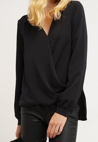 Svart blus med lång ärm, djup V-ringning och veckdetalj. Mjuk tyg, tillsammans med åtsittande svarta byxor.