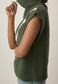 Osoba v bezrukávovém zeleném pleteném svetru s rolákem a bílým pruhem, v kombinaci se světle modrými džíny, se zvednutou paží blízko obličeje.