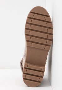 Semelle d'une botte d'hiver beige avec de profondes rainures horizontales et le numéro de taille 37 embossé sur la semelle, positionnée debout contre des murs blancs.