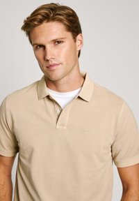 Bež majica s kratkim rukavima od mekog materijala, koja ima klasični ovratnik, placket s tri gumba, izvezeni logo.