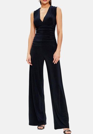 Femme portant une combinaison noire sans manches avec un décolleté en V profond et un pantalon à jambes larges, associée à des talons ouverts noirs, debout devant un fond blanc.