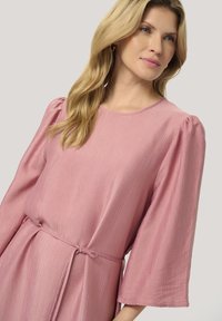Rosa klänning med texturerat tyg, ballongsärmar och knytmidja. Har en rund halsringning och subtila vertikala linjer över materialet.