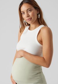 Top de alças branco canelado combinado com uma saia canelada verde-clara. A mão da modelo repousa sobre a barriga, revelando um design justo e elástico.