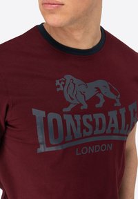 Vyniojamoji trumpomis rankovėmis burgundinė marškinė su laivapriekio mėlyna apykakle ir dideliu pilku liūto grafiniu atvaizdu su tekstu "LONSDALE LONDON" apačioje. Pagaminta iš medvilnės.
