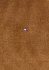 Bruine corduroy stof met verticale ribbels, met een klein geborduurd logo in rood, wit en blauw nabij het midden.