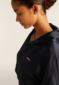 Sudadera con cremallera negra de textura suave, con un logo dorado, capucha con cordón y un corte ajustado.