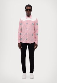 Rosa Langarmhemd mit floralem Stickereid in Blau und Grün, weißer Passe und Kragen, kombiniert mit schwarzen Hosen und weißen Sneakers.