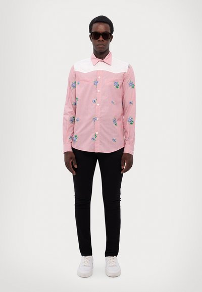 Chemise à manches longues rose avec broderie florale en bleu et vert, empiècement et col blancs, associée à un pantalon noir et des baskets blanches.