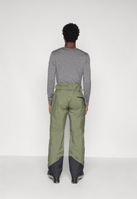 Žali nepralaidūs sniego kelnės su plačiu kirpimu, akcentuoti tamsiai mėlynomis apatinėmis dalimis ir reguliuojamu juosmeniu. Dėvimos virš pilkos ilgomis rankovėmis marškinėlių.