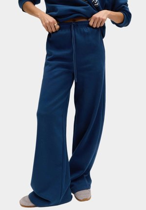 Person iført marineblå vidde sweatpants med snøre i taljen og grå slip-on sko, hænder justerer taljebåndet.