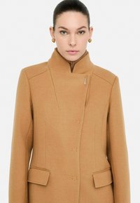Cappotto in misto lana color cammello con colletto alto, linee pulite, tasche laterali e dettagli di cucitura discreti. Presenta una silhouette aderente e hardware minimale.