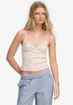 Ung kvinde med langt blondt hår iført en lyserød undertrøje og løstsiddende blåstribede bukser, stående foran en ensfarvet hvid baggrund.