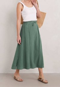 Gonna verde, strutturata, a lunghezza media con vita elastica e dettaglio con fiocco, abbinata a una canotta bianca e sandali beige con fascia.