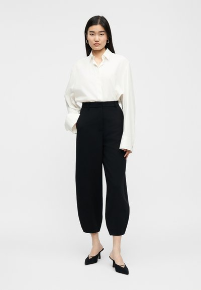 Γυναίκα όρθια με ένα λευκό, φαρδύ πουκάμισο με κουμπιά, μαύρο παντελόνι ψηλόμεσο, φαρδύ, μαύρες μυτερές γόβες και χρυσούς κρίκους σκουλαρίκια.