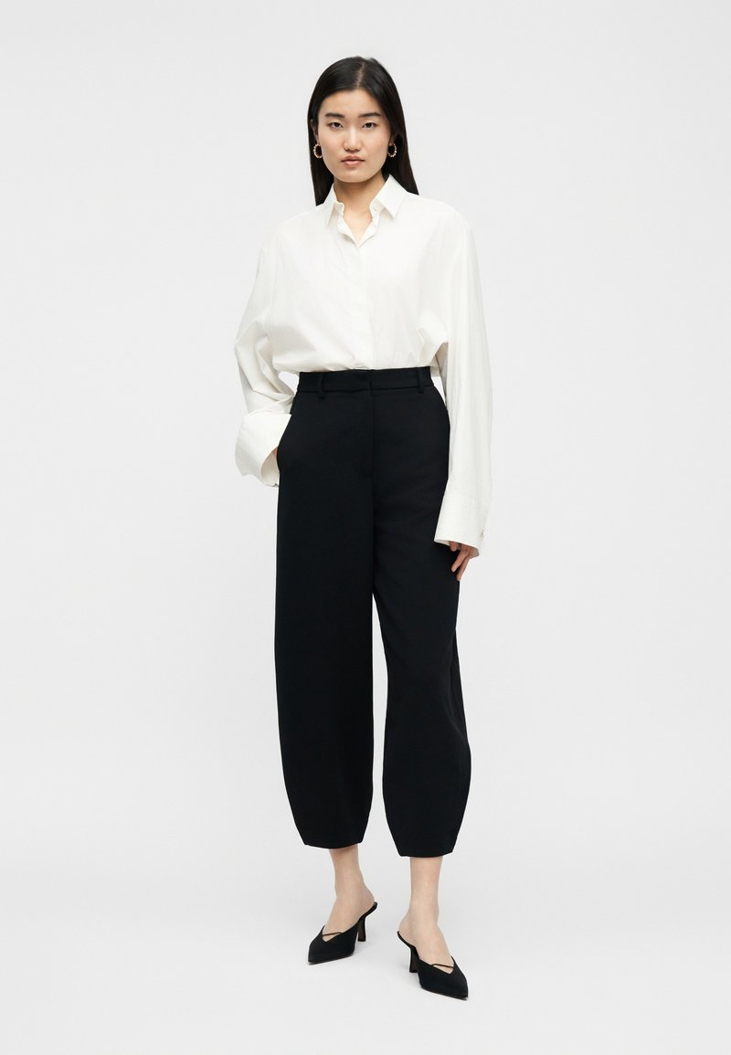 Frau steht da und trägt ein weißes, übergroßes Hemd mit Knöpfen, schwarze, hoch taillierte, weit geschnittene Hosen, schwarze spitze High Heels und goldene Creolen.