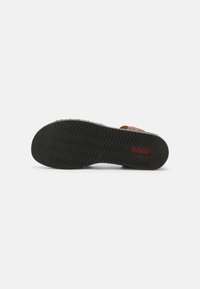 Sandal marron avec une semelle texturée, présentant des motifs de crampons en zigzag pour l'adhérence, et un logo rouge prominent sur la semelle extérieure pour le branding.