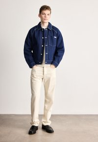 Denimjakke i mørk blå med kontrasterende sømmer, frontlomme og skinnende knapper, kombinert med off-white bukser og svarte sko.