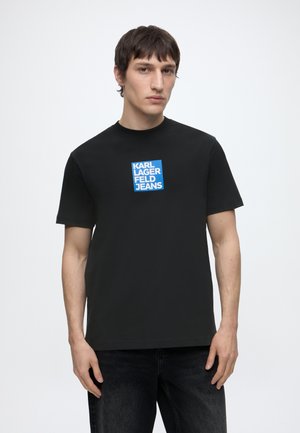 Jeune homme aux cheveux courts et foncés portant un t-shirt noir avec un logo carré bleu indiquant "KARL LAGERFELD JEANS" sur un fond blanc uni.