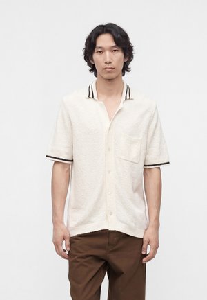 Uomo con capelli neri ondulati che indossa una camicia bianca a maniche corte con bordi neri e pantaloni marroni, in piedi davanti a uno sfondo semplice.