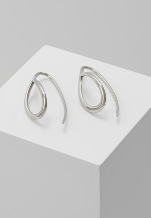 Paire de boucles d'oreilles créoles argentées en forme de larme torsadée reposant sur un cube blanc sur fond gris.