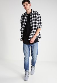 Camisa de cuadros en blanco y negro sobre una camiseta negra, combinada con jeans de mezclilla claros y zapatillas grises. Corte recto y diseño casual.