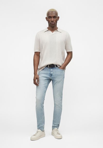 Άνδρας με κοντά ξανθά μαλλιά που φοράει μπεζ κοντομάνικο πλεκτό πόλο μπλουζάκι, ανοιχτό μπλε τζιν παντελόνι, μαύρη ζώνη και λευκά αθλητικά, στέκεται με το ένα χέρι στην τσέπη.